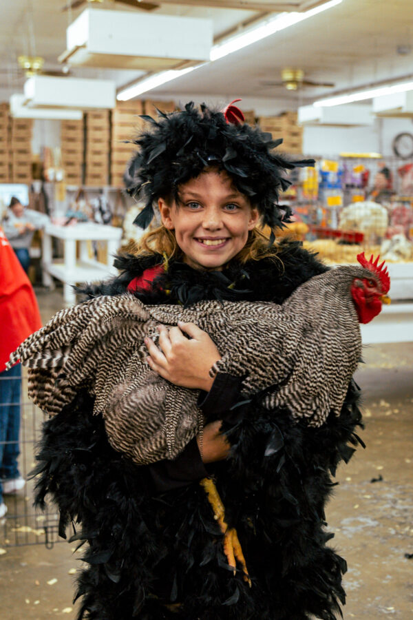 Smiling girl in a black feathered costume holds a large barred chicken in an indoor market or fair setting.